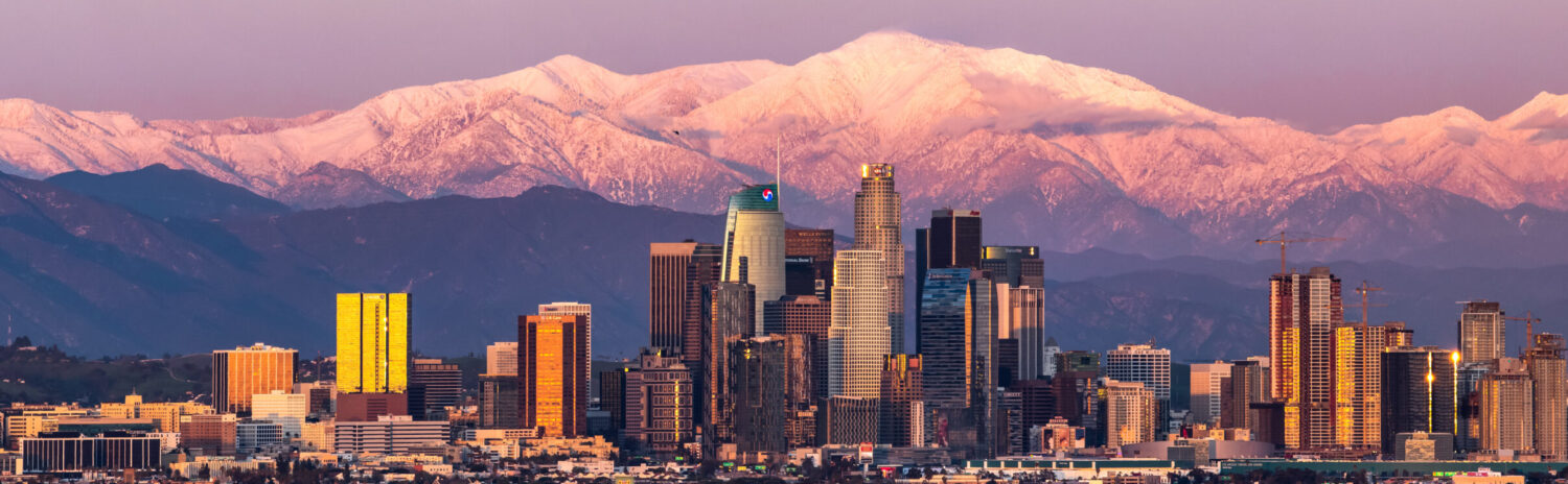 The skyline of Downtown Los Angeles at sunset, with the snow-capped San Gabriel Mountains visible behind it.